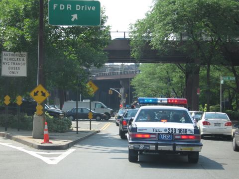1991 Ford LTD Crown Victoria - black-and-white police-style sedan marked TEL. 239-1616 leads a line of cars toward a green FDR Drive ramp sign under an overpass.