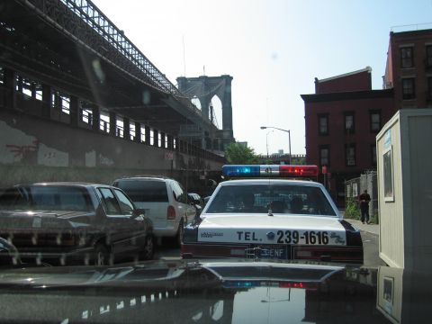 1991 Ford LTD Crown Victoria - black-and-white police-style sedan marked TEL. 239-1616 sits among parked cars on a lot beneath the Brooklyn Bridge and a brick building.
