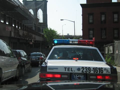 1991 Ford LTD Crown Victoria - white sedan police cruiser with a roof light bar and TEL. 239-1616 lettering sits in traffic beneath the Brooklyn Bridge.