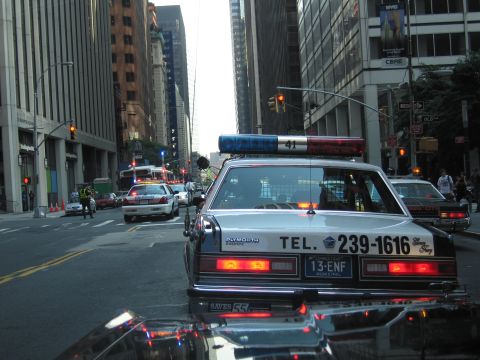 1991 Ford LTD Crown Victoria - dark Plymouth police cruiser marked TEL. 239-1616 follows another patrol car down a city avenue lined with skyscrapers.