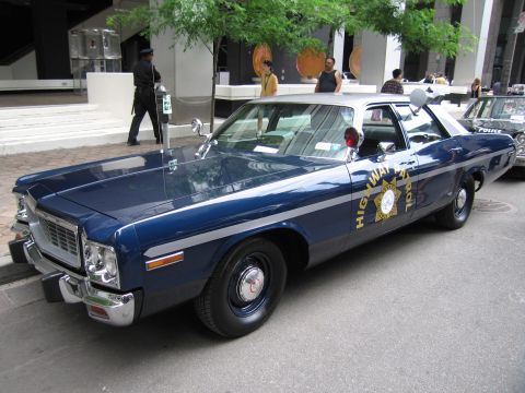1991 Ford LTD Crown Victoria - blue-and-white 1970s Dodge highway patrol sedan with chrome grille and spotlight sits curbside as pedestrians pass behind.