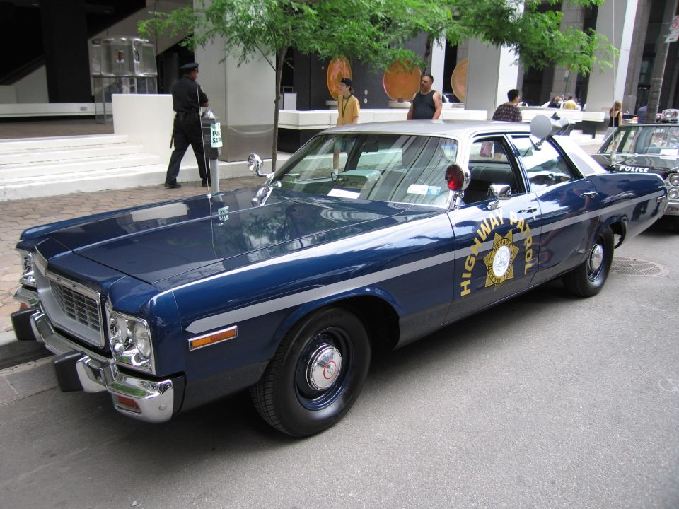 Front three-quarter of blue-and-white 1970s Dodge highway patrol sedan, pedestrians behind