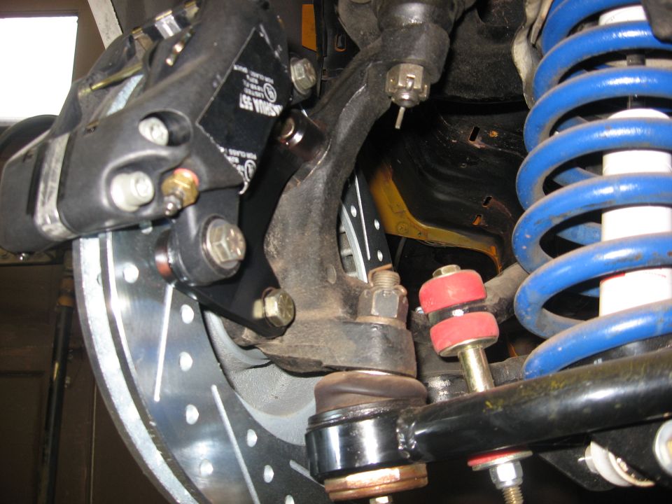 Close-up of a vehicle's front suspension with brake rotor, caliper, blue coil spring, and red bushings.