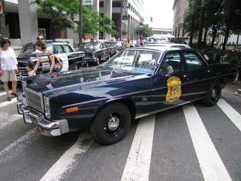 1991 Ford LTD Crown Victoria - dark blue 1970s Dodge Coronet police sedan with gold star door emblem parked over a crosswalk as spectators look on.