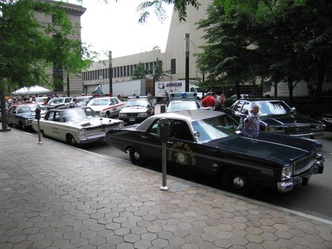 1991 Ford LTD Crown Victoria - lineup of vintage police cruisers parked along a downtown curb, led by a black-and-white highway patrol sedan with star emblem.