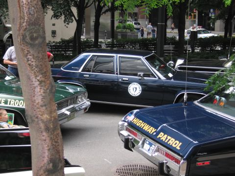 1991 Ford LTD Crown Victoria - dark blue state police sedan with a round seal emblem parked beside a black HIGHWAY PATROL cruiser at a tree-lined car show.
