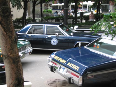 1991 Ford LTD Crown Victoria - dark blue state police sedan with seal emblem and a black HIGHWAY PATROL cruiser parked among trees with city buses in the background.
