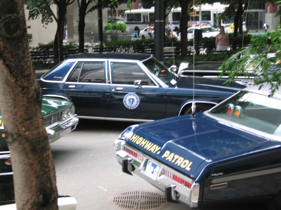 Dark blue state police sedan and black HIGHWAY PATROL car parked among trees, buses behind