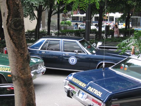 1991 Ford LTD Crown Victoria - dark blue state police sedan with round seal emblem flanked by a green trooper car and a black HIGHWAY PATROL cruiser at a car show.
