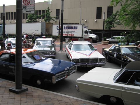 1991 Ford LTD Crown Victoria - Several restored vintage police cruisers, one dark blue and one a white striped wagon, are parked tightly together on a city street.