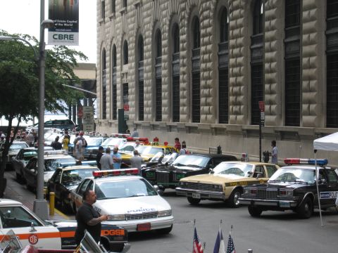 1991 Ford LTD Crown Victoria - People walk among rows of classic and modern police cars displayed beside a stone building on a city street.