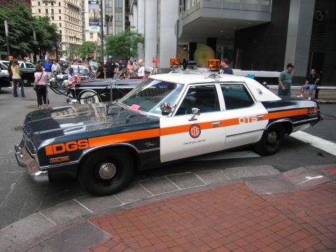 1991 Ford LTD Crown Victoria - vintage DGS official sedan in blue and orange livery sits on a brick plaza as a crowd browses other cars behind it.