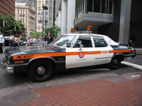 1991 Ford LTD Crown Victoria - side of a vintage DGS official sedan with blue and orange striping and a roof light is parked on a city brick plaza.