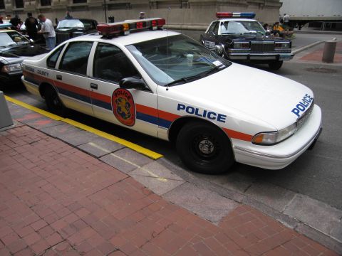 1991 Ford LTD Crown Victoria - white Nassau County police sedan with a roof light bar and orange seal decal is parked at the curb near other cruisers.