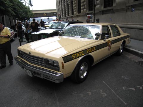 1991 Ford LTD Crown Victoria - tan and black Chevrolet State Police coupe with a roof light is displayed on a city street among other police cars.