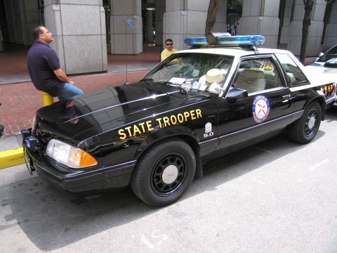 1991 Ford LTD Crown Victoria - black Ford Mustang 5.0 marked State Trooper with a roof light bar sits on a city street as a man leans on a planter nearby.