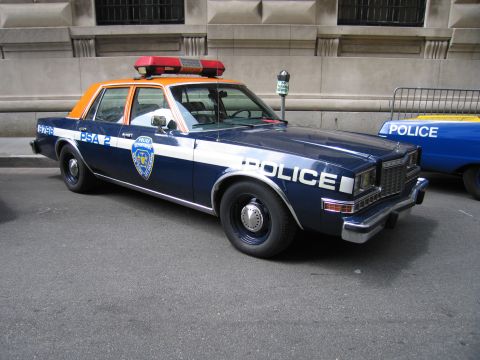 1991 Ford LTD Crown Victoria - vintage blue, white and orange NYPD police sedan with a red roof light bar is parked beside a metal barricade on the street.