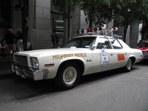 1991 Ford LTD Crown Victoria - cream-colored vintage Highway Patrol sedan with roof spotlights and the number 2250 is parked on a tree-lined city street.