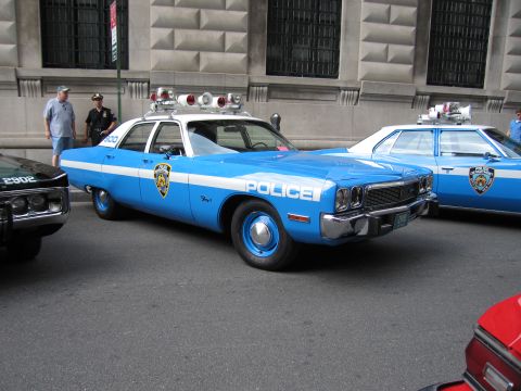 1991 Ford LTD Crown Victoria - light blue and white vintage NYPD police sedan with twin roof lights is parked among other cruisers beside a stone building.