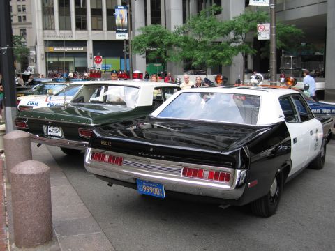 1991 Ford LTD Crown Victoria - rear of two vintage Plymouth police sedans, one black and white and one green, parked at a downtown curb among onlookers.