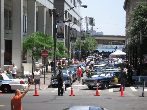 1991 Ford LTD Crown Victoria - sunny downtown street is filled with rows of vintage police cars and crowds of spectators near banners and traffic cones.