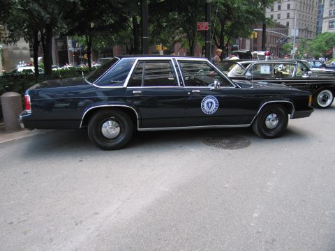 1991 Ford LTD Crown Victoria - dark navy four-door Ford Crown Victoria police sedan bearing a round state seal on the door, parked curbside among trees in a city.