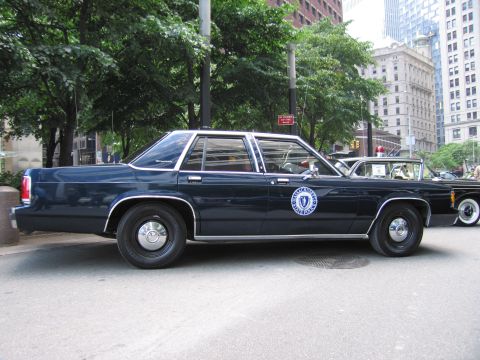 1991 Ford LTD Crown Victoria - Profile of a dark navy Ford Crown Victoria police cruiser with a state seal on the front door, parked on a tree-lined urban street.