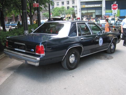 1991 Ford LTD Crown Victoria - Rear three-quarter view of a dark navy Ford police sedan with a round door seal, parked near a Washington Mutual storefront and a stop sign.