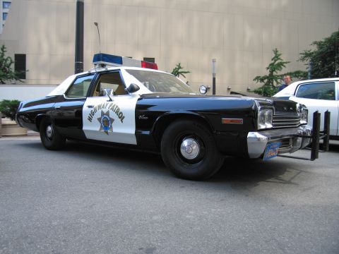 1991 Ford LTD Crown Victoria - black-and-white Dodge Highway Patrol cruiser with star door emblem, front push bar, and roof lightbar parked along a building.