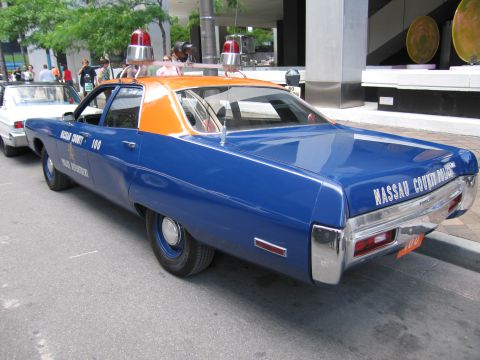 1991 Ford LTD Crown Victoria - Rear three-quarter of a blue Plymouth police sedan with orange roof, twin red beacons, and Nassau County Police Department lettering on the…
