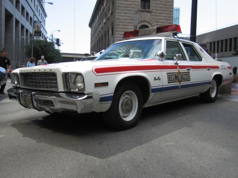 1991 Ford LTD Crown Victoria - white Plymouth State Police sedan with red and blue body stripes, a star door emblem, and roof lightbar parked on a city street.