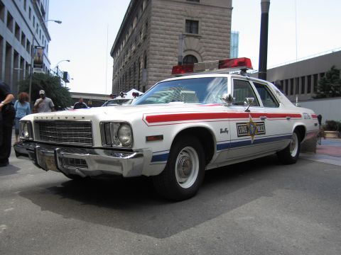 1991 Ford LTD Crown Victoria - Front three-quarter of a white Plymouth State Police sedan with red and blue stripes, star door emblem, and roof lights among city building…
