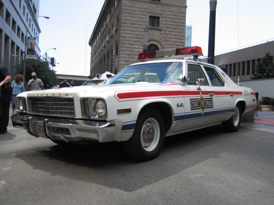 White Plymouth State Police cruiser with red-white-blue stripes and front push bar