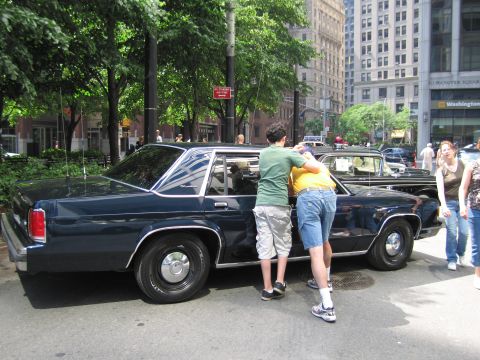 1991 Ford LTD Crown Victoria - Two young people lean into the open driver's door of a dark navy Ford sedan parked curbside, with pedestrians and buildings behind.