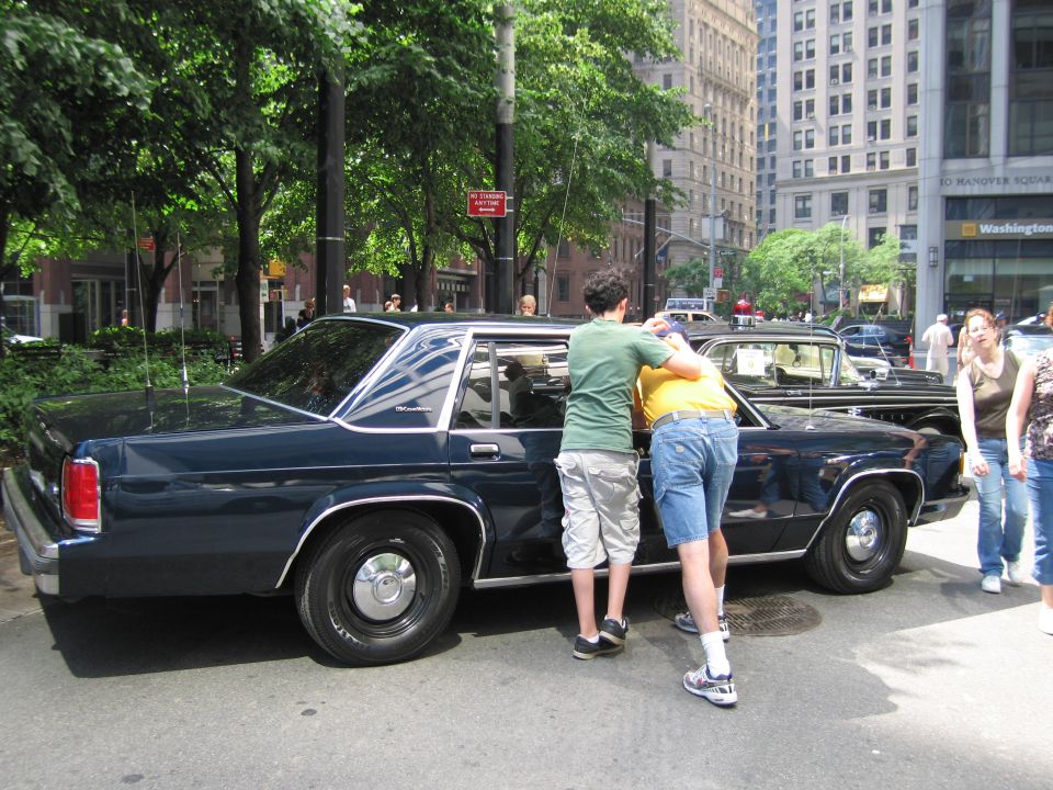 Two people lean into open door of dark blue Ford sedan on city street