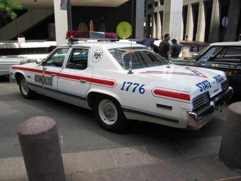 1991 Ford LTD Crown Victoria - Rear three-quarter of a white State Police sedan with patriotic stripes, an eagle emblem, and the number 1776 on the rear door, by a plaza.