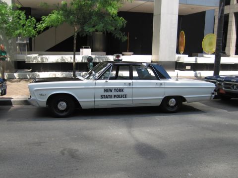 1991 Ford LTD Crown Victoria - Profile of a white Plymouth sedan lettered New York State Police with a single roof beacon, parked on a tree-lined city street.