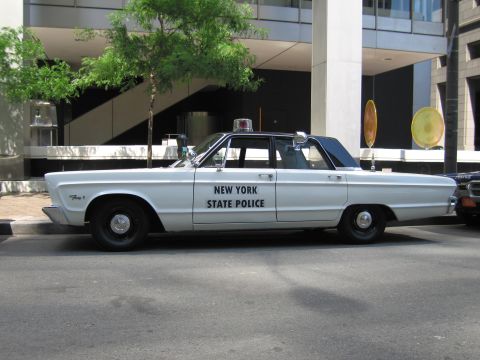 1991 Ford LTD Crown Victoria - Side view of a white Plymouth sedan marked New York State Police with a single roof beacon, parked in front of a modern building.