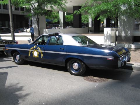 1991 Ford LTD Crown Victoria - Profile of a dark blue Highway Patrol sedan with a white roof, roof beacon, and gold star door emblem parked along a city curb.