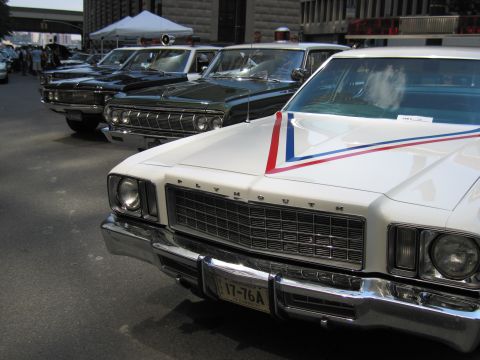 1991 Ford LTD Crown Victoria - Close front view of a white Plymouth police sedan with patriotic hood stripe and a 17-76A plate, parked in a row of classic cars.