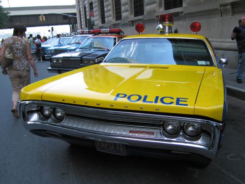 1991 Ford LTD Crown Victoria - Front view of a bright yellow Plymouth sedan lettered POLICE in blue with twin red roof beacons, parked on a city street as a woman walks p…