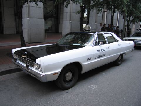 1991 Ford LTD Crown Victoria - white Plymouth sedan with a black hood lettered New York State Police, a single roof beacon, parked beside a stone building.