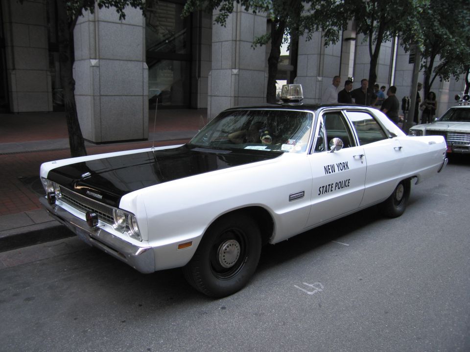 White Plymouth New York State Police sedan with black hood, parked by stone building