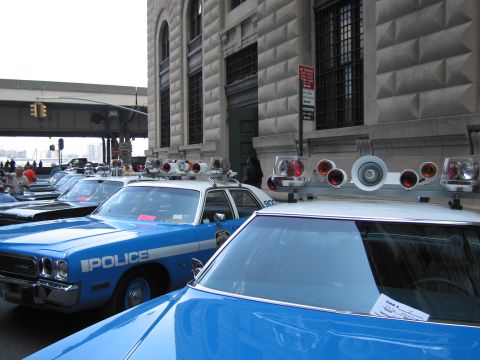 1991 Ford LTD Crown Victoria - blue Dodge sedan lettered POLICE parked beside another blue car whose roof carries a cluster of red and white emergency lights, by a buildi…