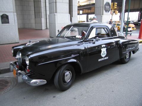 1991 Ford LTD Crown Victoria - black Studebaker coupe with a bullet nose lettered NYPD Detective Bureau, with an NYPD shield on the door, parked on a city street.