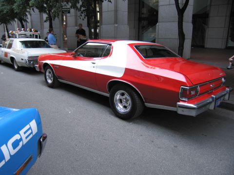 1991 Ford LTD Crown Victoria - red two-door coupe with a wide white vector stripe, evoking the Starsky and Hutch car, parked at a city curb beside police vehicles.