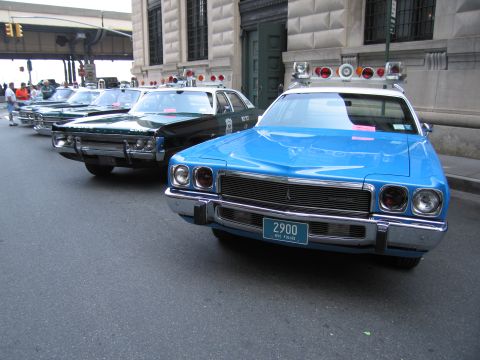 1991 Ford LTD Crown Victoria - blue vintage police sedan with roof lightbar and plate 2900 NYC Police leads a row of classic cruisers along a city building.