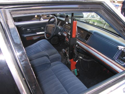 1991 Ford LTD Crown Victoria - Interior of a vintage police sedan showing blue cloth bench seats, woodgrain dash and an orange-stocked shotgun in a center mount.