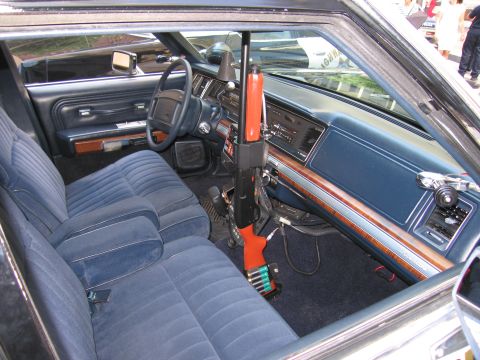 1991 Ford LTD Crown Victoria - police sedan interior with blue velour bench seat, woodgrain dashboard, radio gear and an orange-stocked shotgun upright in a mount.