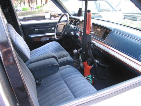 1991 Ford LTD Crown Victoria - Through an open door, a vintage police car shows a blue cloth bench seat, woodgrain dash and a mounted orange-stocked shotgun.
